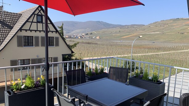 Un balcón con una mesa y sillas, una sombrilla roja y vistas a un viñedo y colinas.