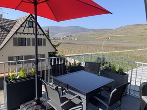 Ein Balkon mit Tisch und Stühlen, einem roten Sonnenschirm und Blick auf einen Weinberg und Hügel.