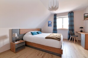 Ein modernes Schlafzimmer mit Bett, Nachttisch, Lampe, Sessel, Schreibtisch und einem Fenster mit Vorhängen.