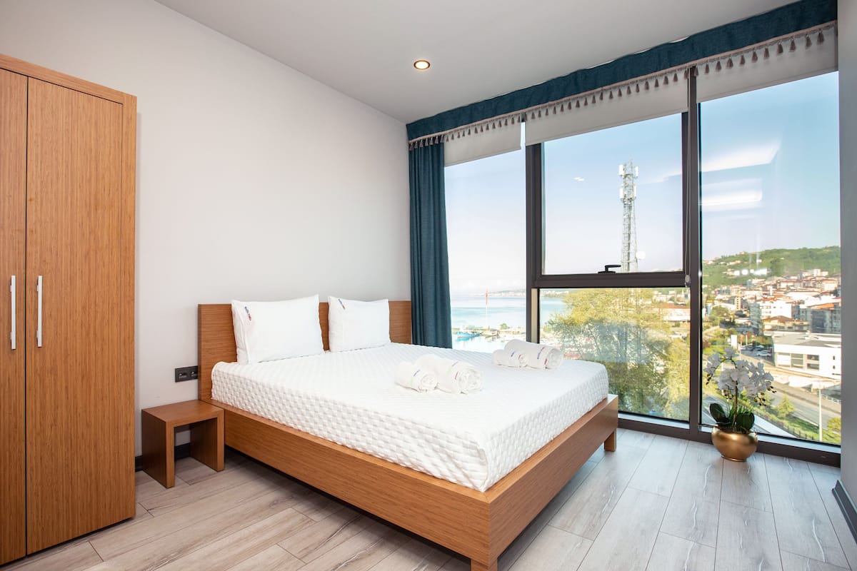 Un dormitorio con una cama grande, un armario de madera, una mesita de noche pequeña y vistas a la ciudad a través de amplios ventanales.