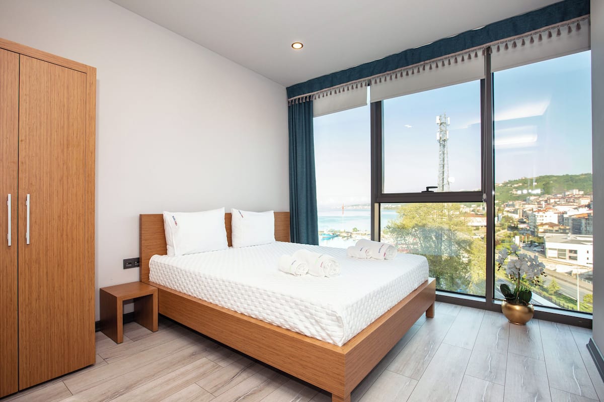 Un dormitorio moderno con una cama grande, un armario de madera y vistas a un paisaje urbano a través de un ventanal.