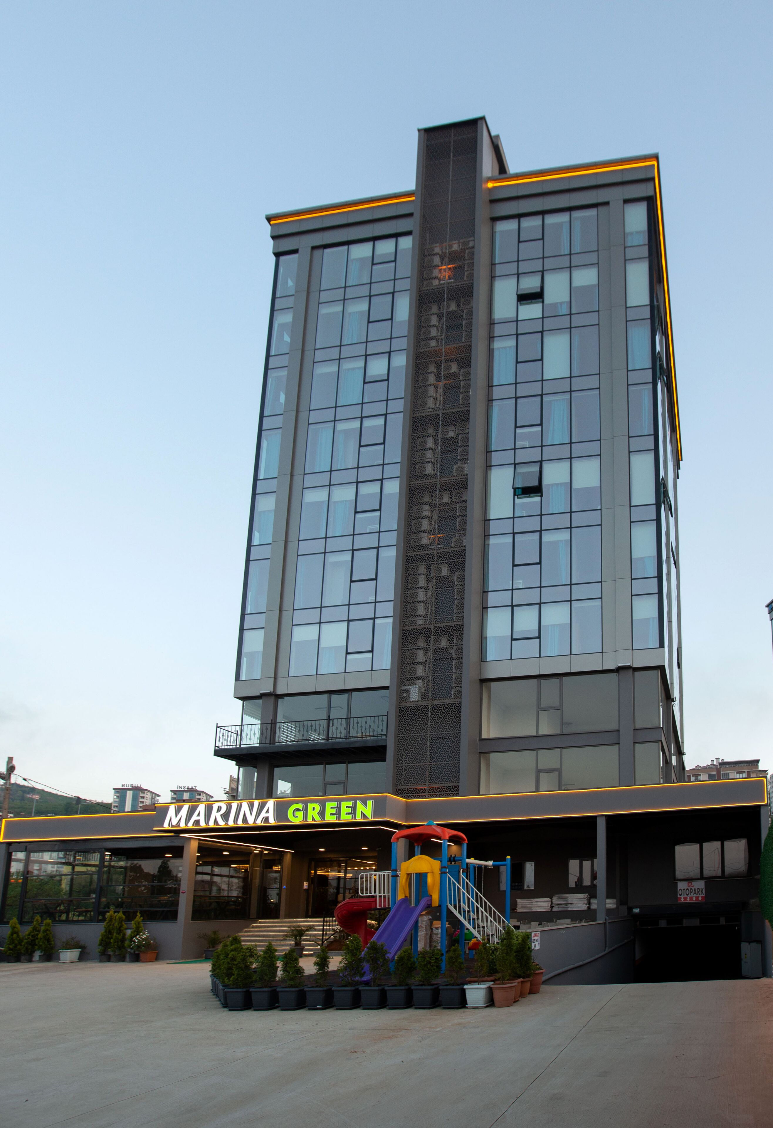 Un edificio moderno con un gran frente de vidrio, entrada visible y una zona de juegos infantiles con toboganes y escaleras.