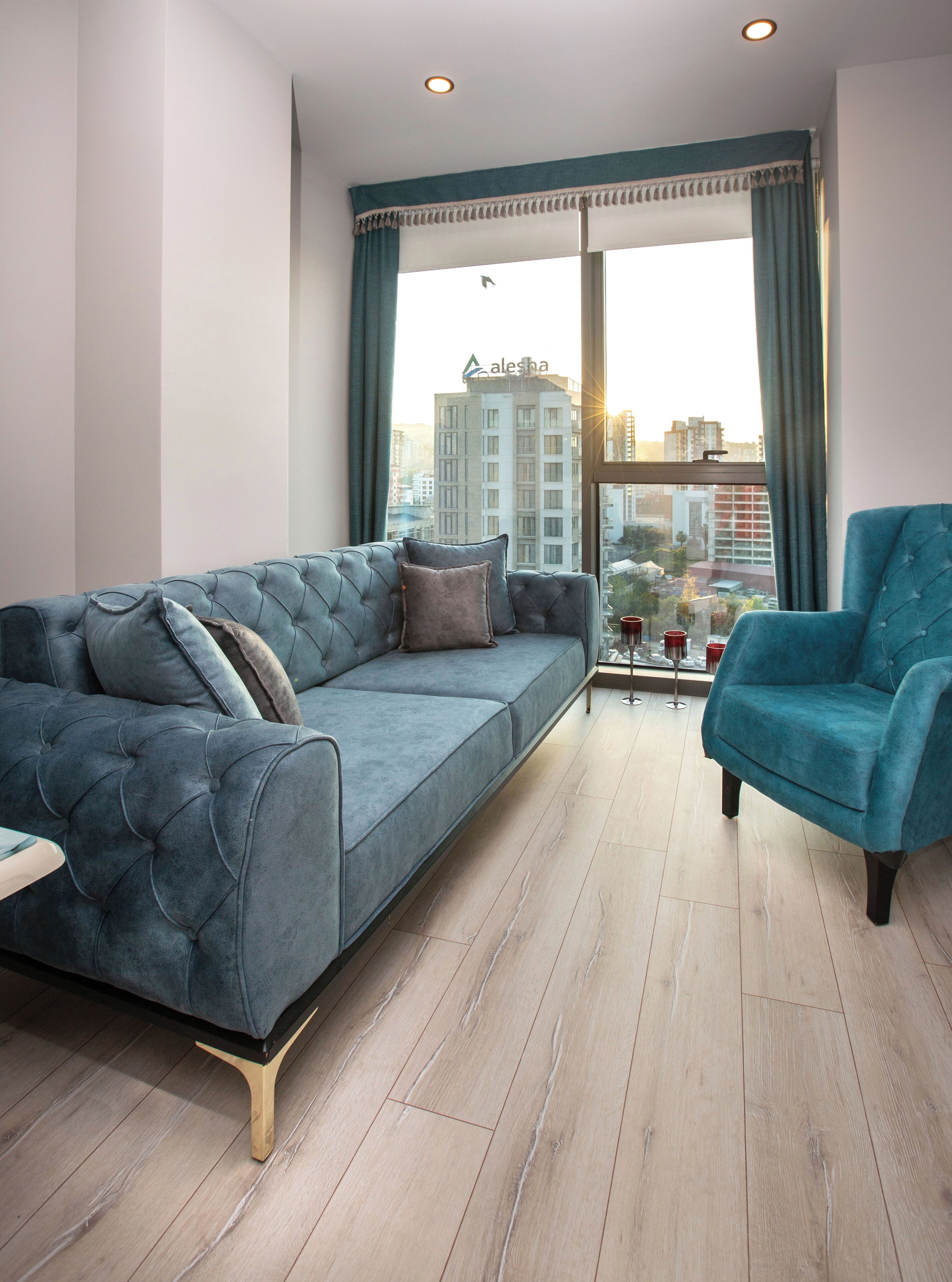 A modern living room with a grey sofa and a teal armchair, wooden flooring, and a large window with curtains.
