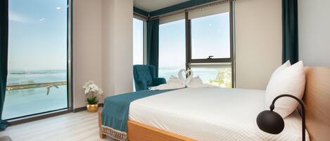 A bedroom with a large bed, a chair, a desk, and a view of the sea.