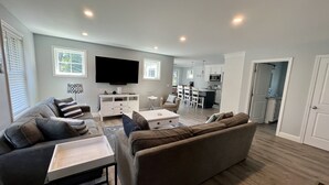 A modern living room with a sofa, a coffee table, and a TV. There is a kitchen in the background with white cabinets and a dining area.