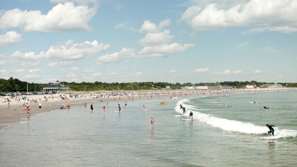 A crowded beach with people swimming and surfing, and a clear sky.