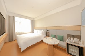 A hotel room with a bed, a round table with a tray of items, a minibar, and a large window with curtains.