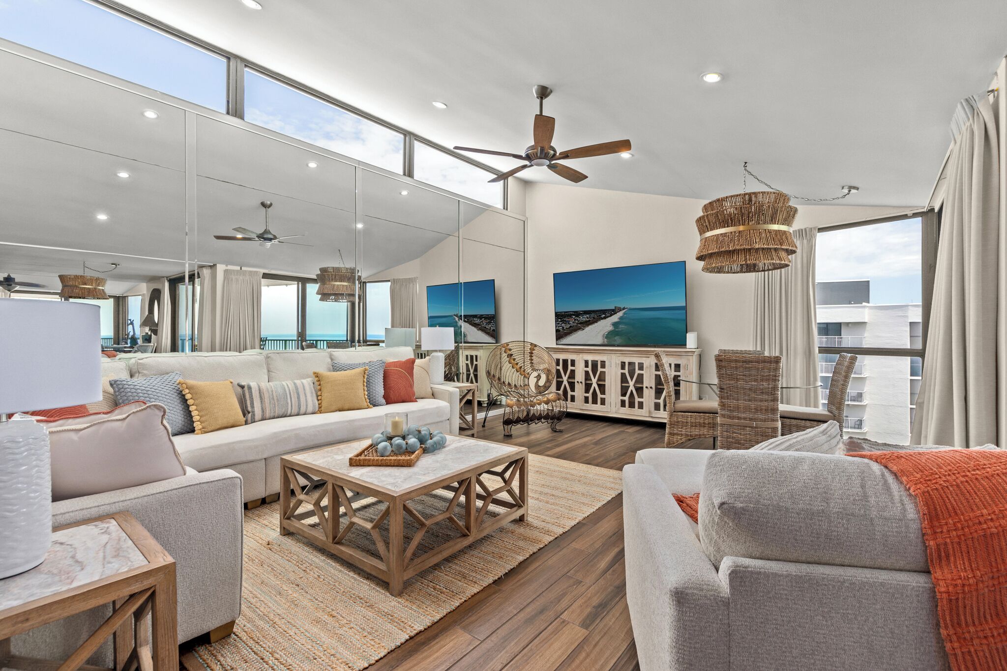 A spacious living room with a large sofa, a coffee table, and a dining area. There are wicker chairs and a ceiling fan. The room has large windows with a view of the ocean.