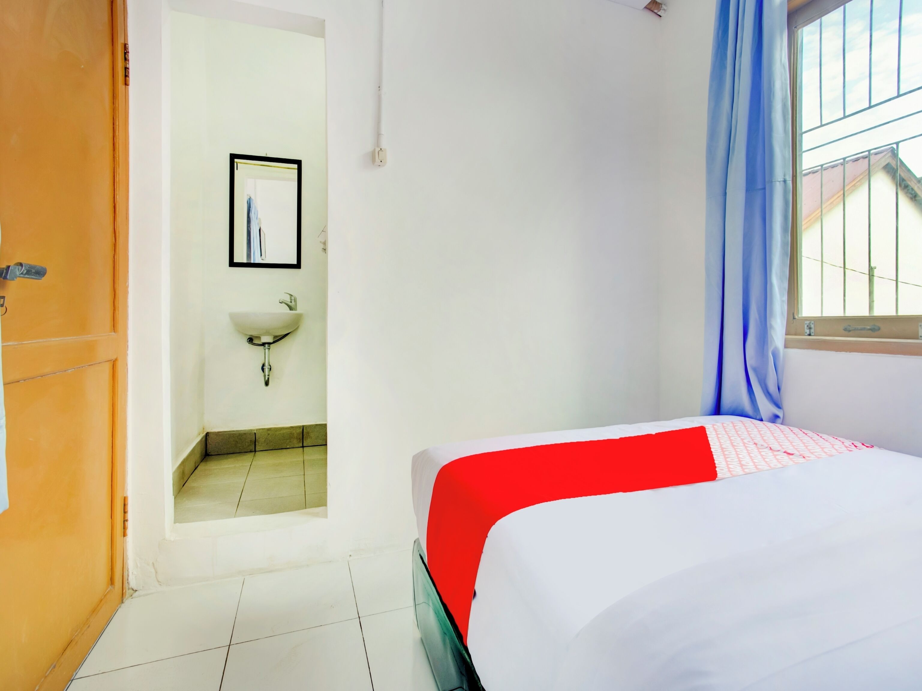 A hotel room with a bed, a window with blue curtains, a sink, and a framed picture on the wall.