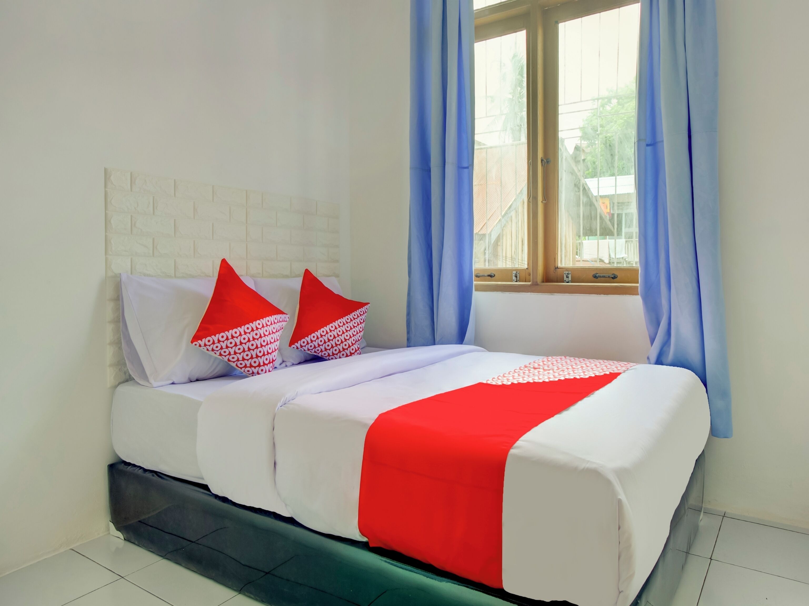 A bed with white bedding and red accents, a window with blue curtains, and a white brick wall.