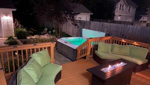 Una terraza de madera con un jacuzzi, dos sillones verdes acolchados y un pozo de fuego.