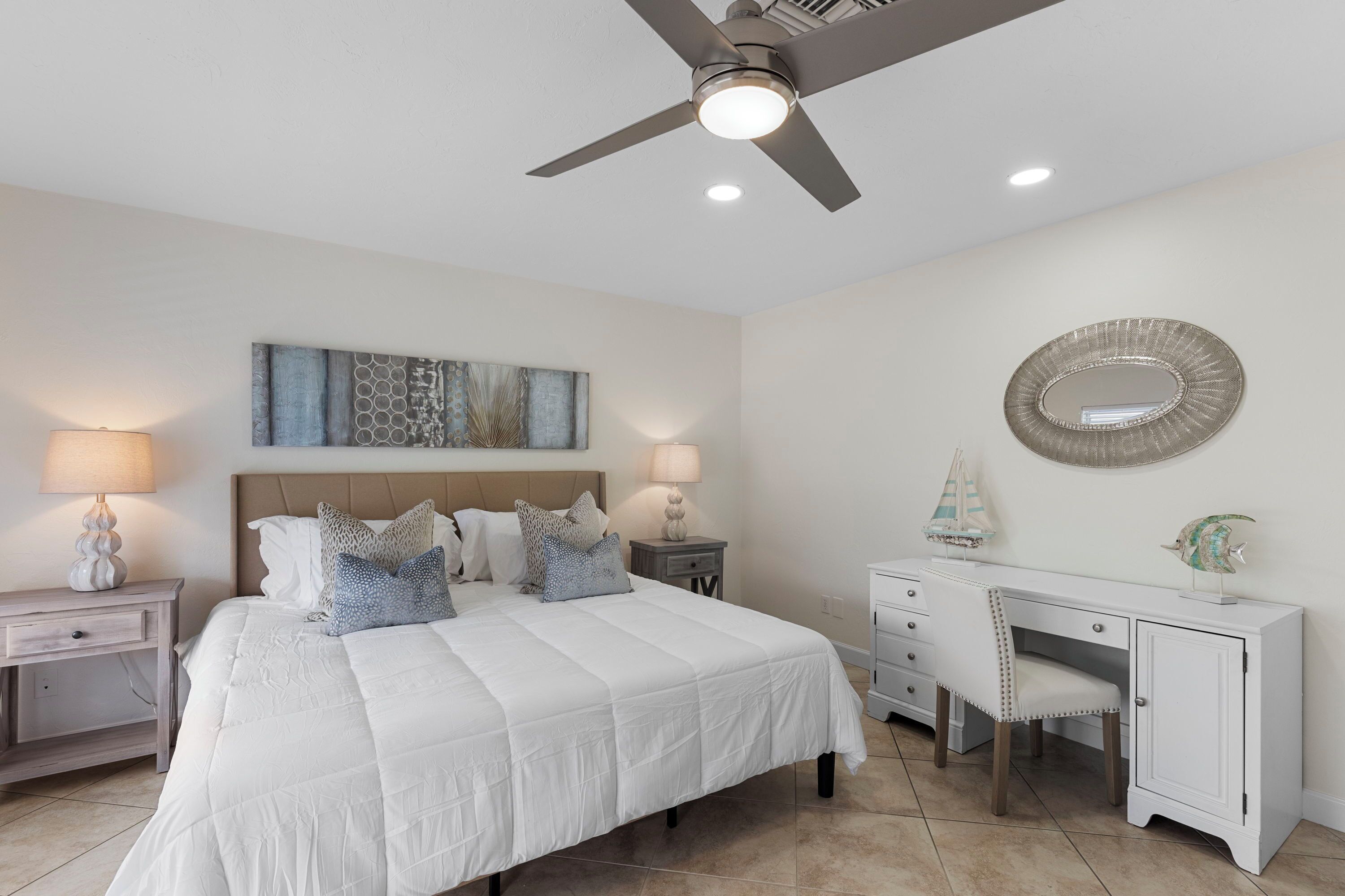 A bedroom with a bed, nightstands, a desk, a chair, a ceiling fan, and a wall-mounted mirror.