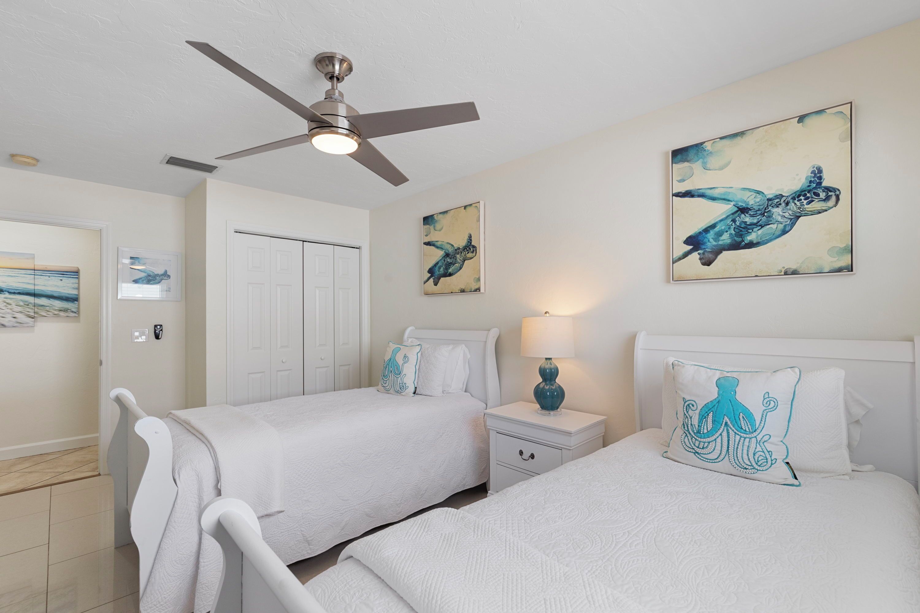 A hotel room with two beds, a ceiling fan, artwork on the walls, and a nightstand with a lamp.