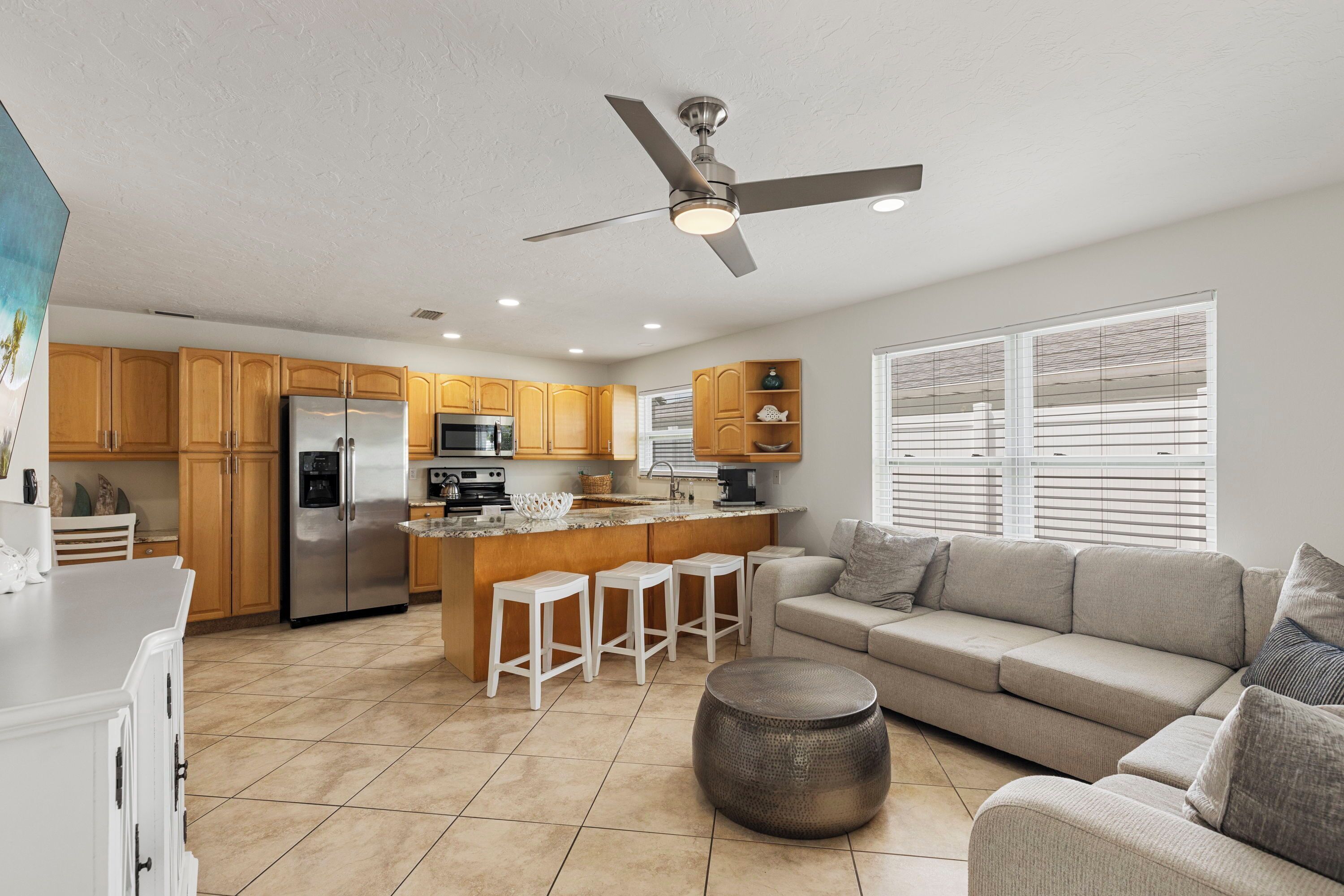 A modern kitchen with wooden cabinets, a stainless steel refrigerator, a built-in island with stools, and a ceiling fan.