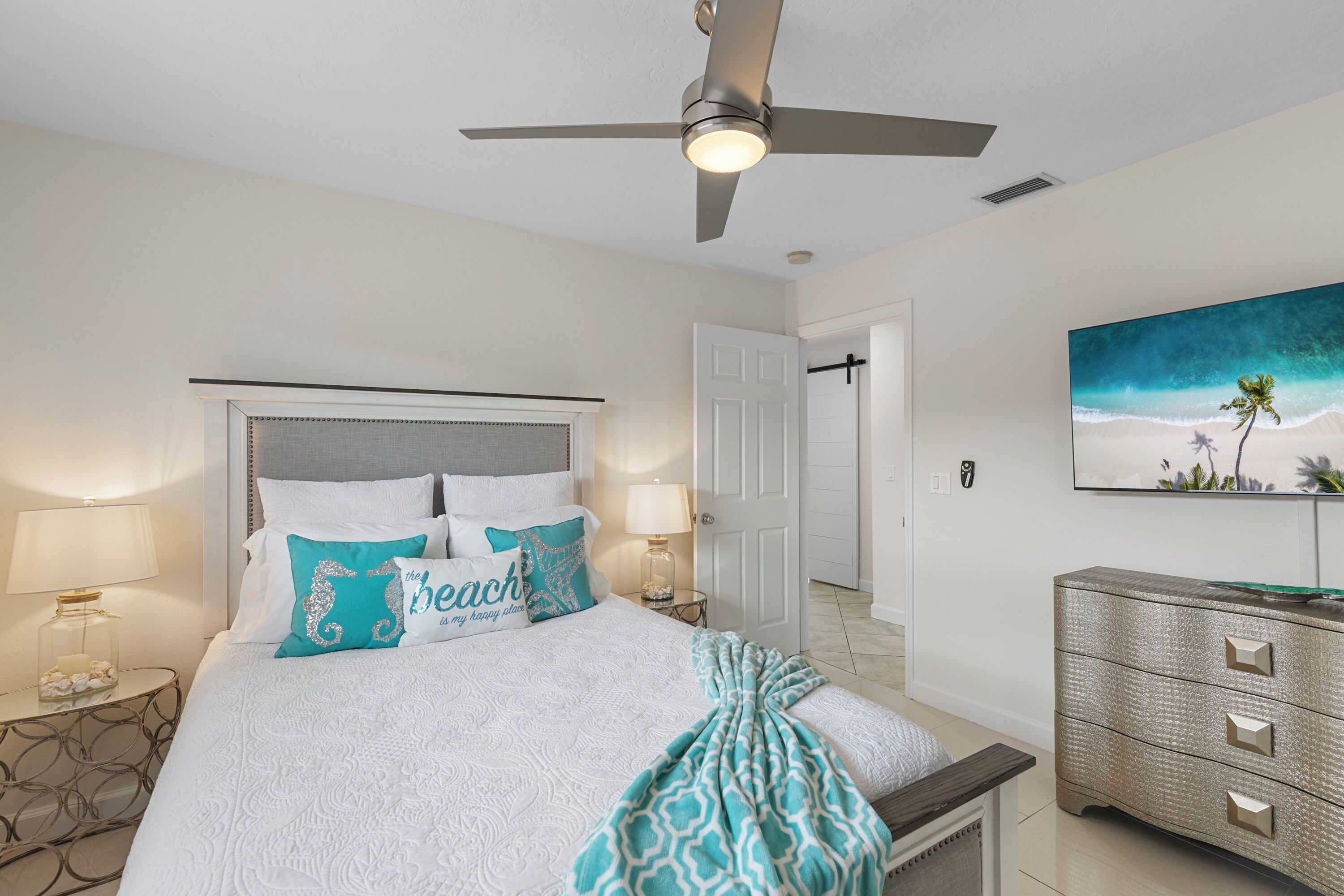 A bedroom with a bed, a ceiling fan, a nightstand with a lamp, a dresser, and a painting on the wall.