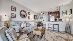 A modern living room with a grey sofa, wooden coffee table, and a kitchen area with a fridge, stove, and bar stools. - Walk To Beach! Beautiful Ocean Views from Condo W/ Lanai, Pool & Hot Tub, BBQ (Kihei)