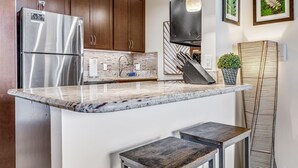 A modern kitchen with a stainless steel refrigerator, wooden cabinets, a marble countertop, and pendant lighting. - Walk To Beach! Beautiful Ocean Views from Condo W/ Lanai, Pool & Hot Tub, BBQ (Kihei)
