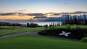 A golf course with a large white flower sculpture, a water body, and a forested area in the background. - Steps from Merriman's, Your Kapalua Villa stay (Lahaina)