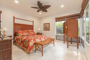 A bedroom with a large bed, a wooden headboard, a ceiling fan, a bedside table with a lamp, a window with blinds, and a view of greenery outside.