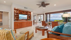 A spacious living room with a large TV, wooden cabinets, wicker furniture, and a view of the ocean. - Steps from Merriman's, Your Kapalua Villa stay (Lahaina)