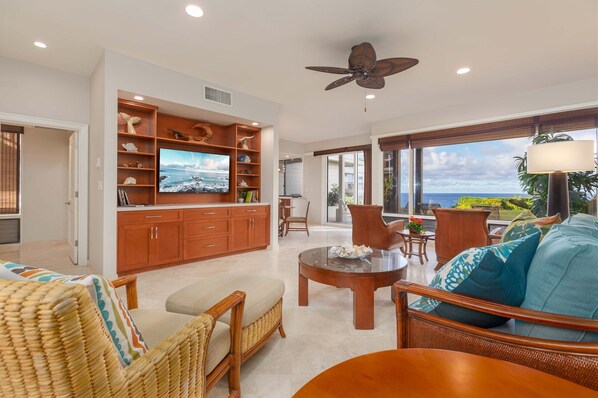A spacious living room with a large TV, wooden cabinets, wicker furniture, and a view of the ocean.