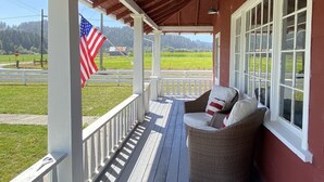 A covered porch with a wicker chair and cushions, white railings, and an American flag. - The Red Farmhouse (Ferndale)