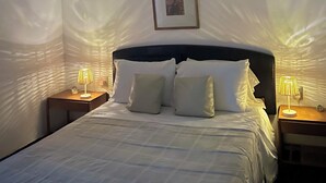 Una cama bien tendida con sábanas blancas, dos mesitas de noche con lámparas, un cuadro enmarcado en la pared y una lámpara colgante.
