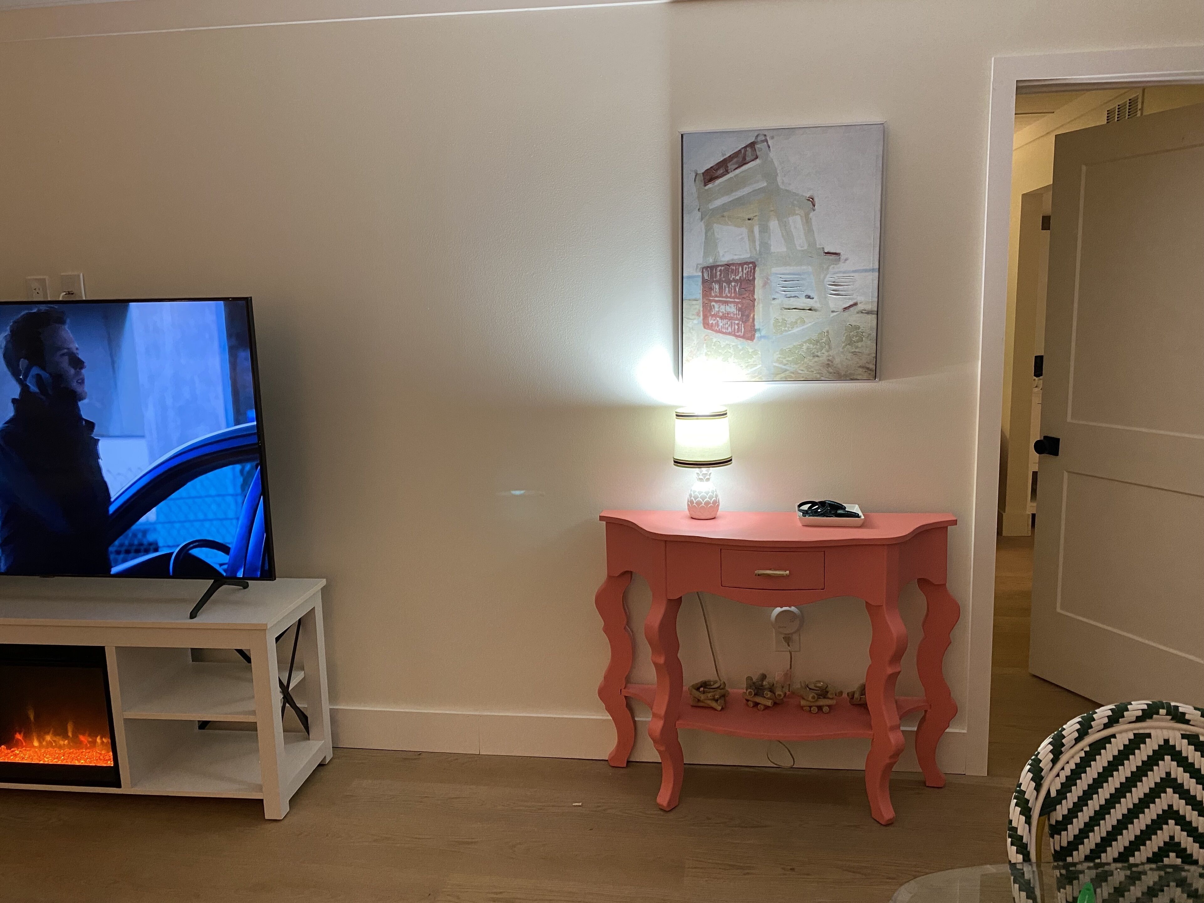 Una sala de estar con televisor, una consola rosa, una lámpara pequeña y un cuadro en la pared.