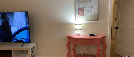 Una sala de estar con televisor, una consola rosa, una lámpara pequeña y un cuadro en la pared.