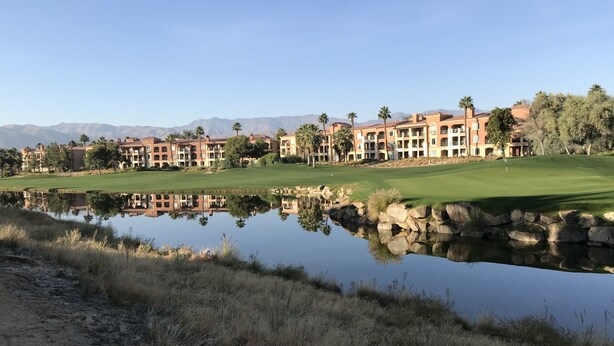 Ein Golfplatz mit Teich, in dem sich Gebäude spiegeln, umgeben von trockenem Gras und Palmen.