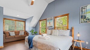 A bedroom with a large bed, a sofa, and a small table with a lamp. There are posters on the wall and a window with blinds.