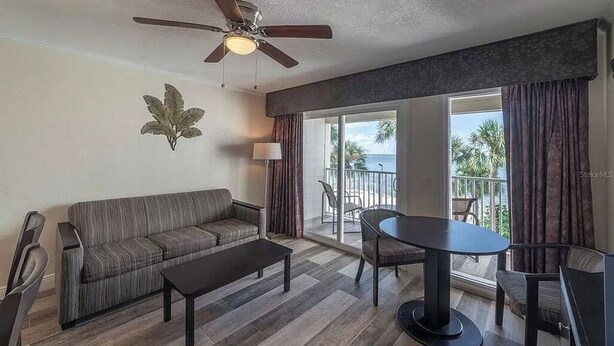 Un salon moderne avec un canapé, une table basse et une table à manger avec des chaises. On peut voir un ventilateur de plafond, et il y a une grande fenêtre avec des rideaux.