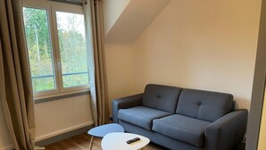 Un petit salon avec un canapé gris, une table basse blanche et une fenêtre avec des rideaux.