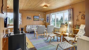 A cozy cabin interior with a wooden ceiling, a sofa, multiple chairs, a table, a fireplace, and a large window offering a view of trees. - Nice home in Holbæk with kitchen (Holbæk)