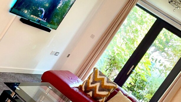 A living room with a flat-screen TV mounted on the wall, a red leather sofa with patterned cushions, a glass coffee table, and a window with a view of greenery. - Affordable luxury apartment (Stratford)