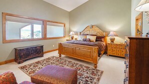 A bedroom with a large bed, a wooden dresser, a nightstand, a mirror, and a window with blinds. - Spacious Mtn Retreat - 3 Minute Walk to Slopes + Hot Tub & Garage (Crested Butte)