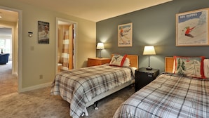 Une chambre avec deux lits, une table de chevet et une lampe. Il y a des affiches sur le mur et une porte qui donne sur une autre pièce.