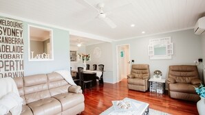 A spacious living room with a wooden floor, a beige sofa, a brown armchair, and a dining area with a table and chairs.