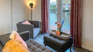 Una sala de estar moderna con un sofá, un sillón y una pequeña mesa con velas y flores. Se ve una lámpara de pie y una vista de un canal con barcos a través de la ventana.