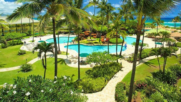 Ett semesterhotell med en central pool omgiven av palmer och en strand.