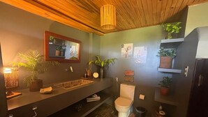 Un baño moderno con techo de madera, un espejo grande, un lavamanos de diseño geométrico, un inodoro y diversas plantas en macetas.