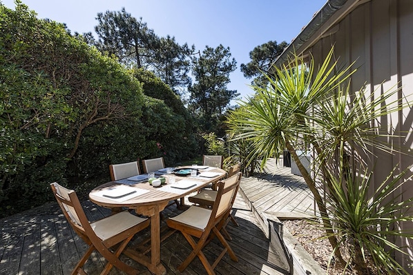 Une terrasse extérieure en bois, avec une table ronde et des chaises, entourée d’une végétation luxuriante et d’un palmier.