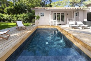A rectangular swimming pool with a wooden deck, surrounded by lounge chairs and a small building with a tiled roof.