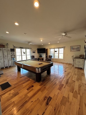 A spacious room with a pool table, wooden flooring, ceiling fans, and a television mounted on the wall.