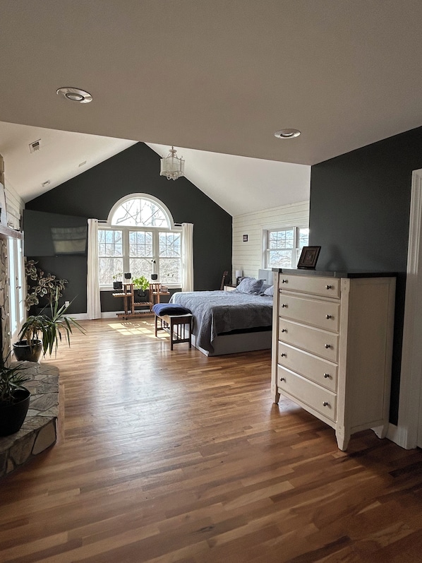 A spacious room with a large bed, a dresser, and a desk with a chair. There is a window with a view of trees and a door leading to another room.