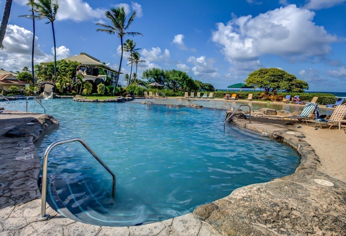 Un área de piscina con agua azul cristalina, una terraza de piedra, sillas de descanso, palmeras y un edificio al fondo.