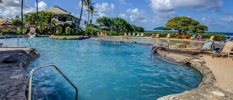 Un área de piscina con agua azul cristalina, una terraza de piedra, sillas de descanso, palmeras y un edificio al fondo.