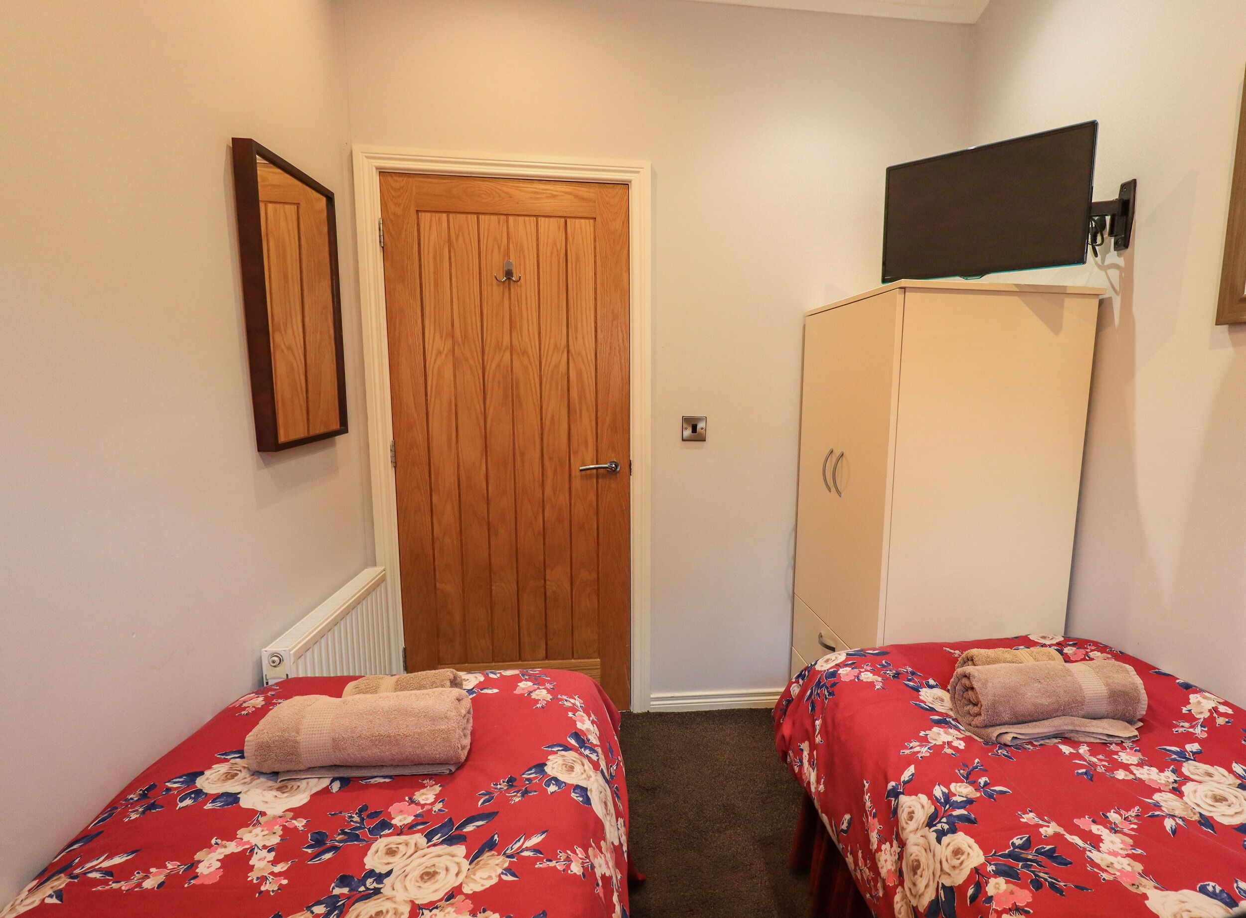 A bedroom with two single beds, a wooden wardrobe, a television mounted on the wall, and a door with a handle.