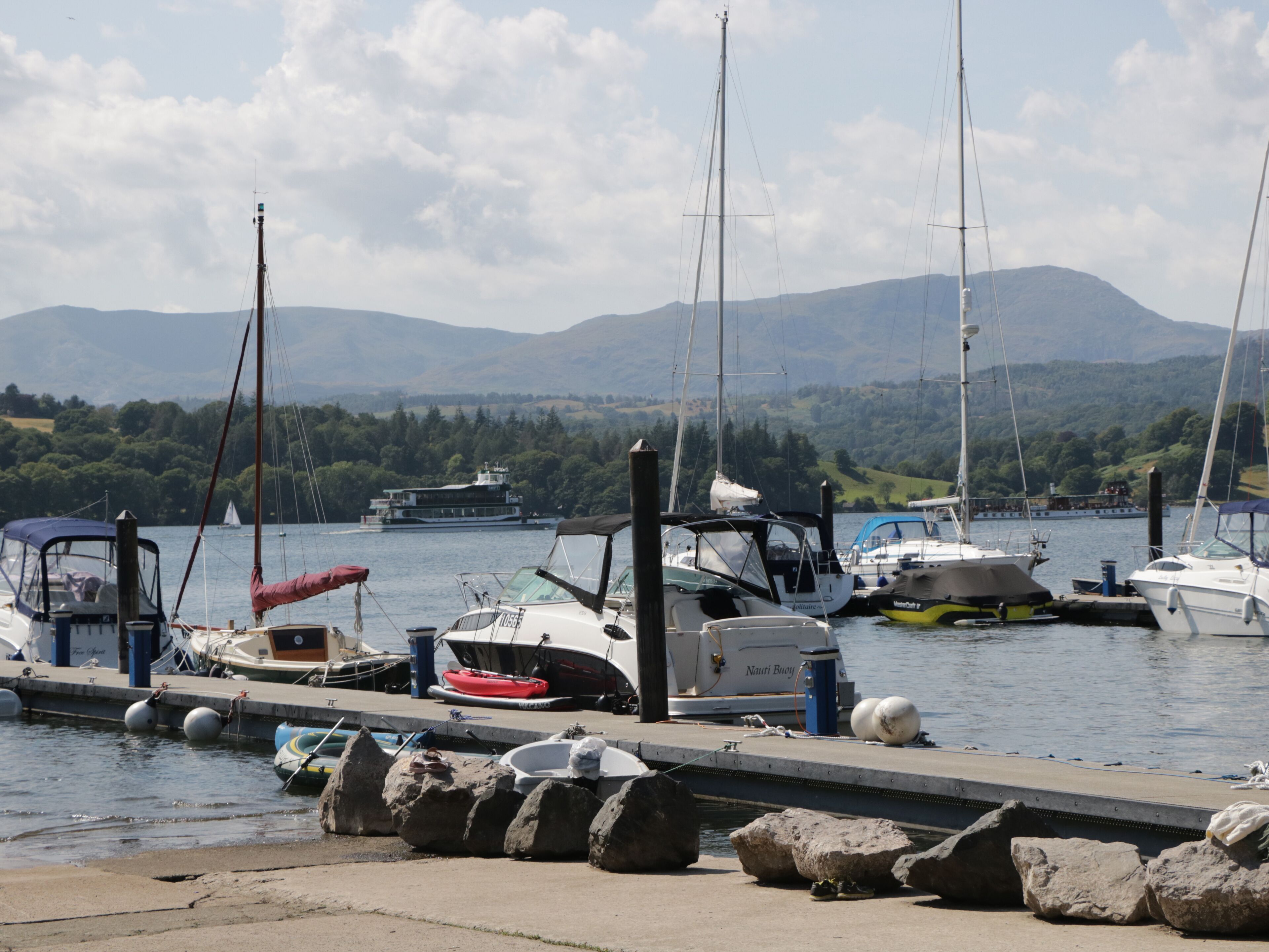 A marina with boats docked, a rocky breakwater, and mountains in the background.