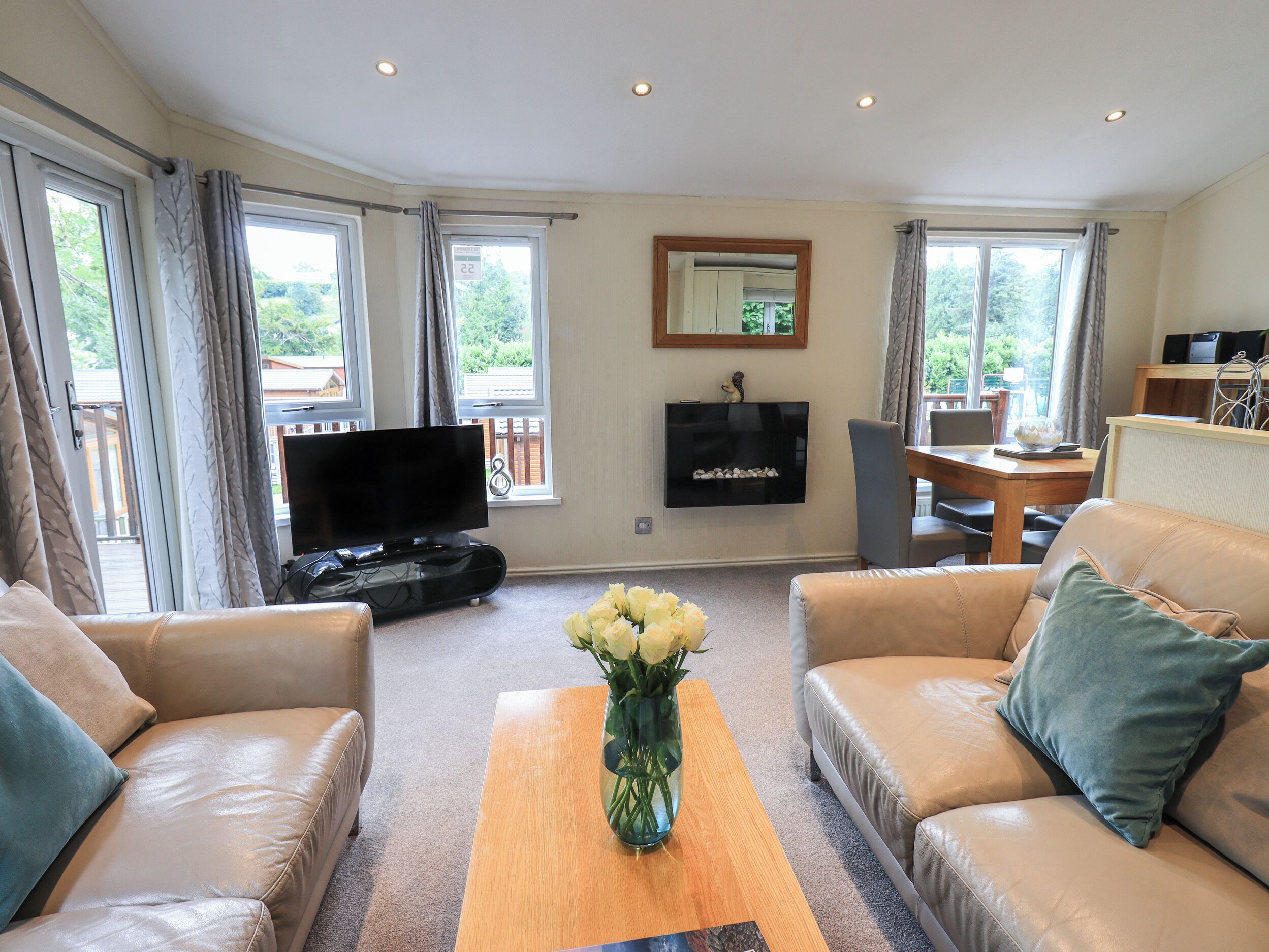 A living room with a flat-screen TV, leather sofas, a coffee table with a vase of flowers, and large windows with curtains.